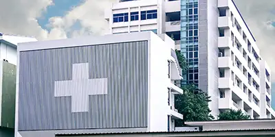Gebäudekomplex mit einem großen, weißen Plus auf der Fassade im Vordergrund. Im Hintergrund ist der weitere Gebäudeteil mit Balkonen und Fenstern zu sehen.