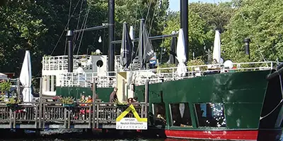 Ein grünes Ausflugsschiff liegt vor Anker am Pier. Das Schiff hat im Bug große Fenster. An Deck stehen Sonnenschirme. Bild zur Verfügung gestellt von Van Loon Restaurantschiffe, Berlin
